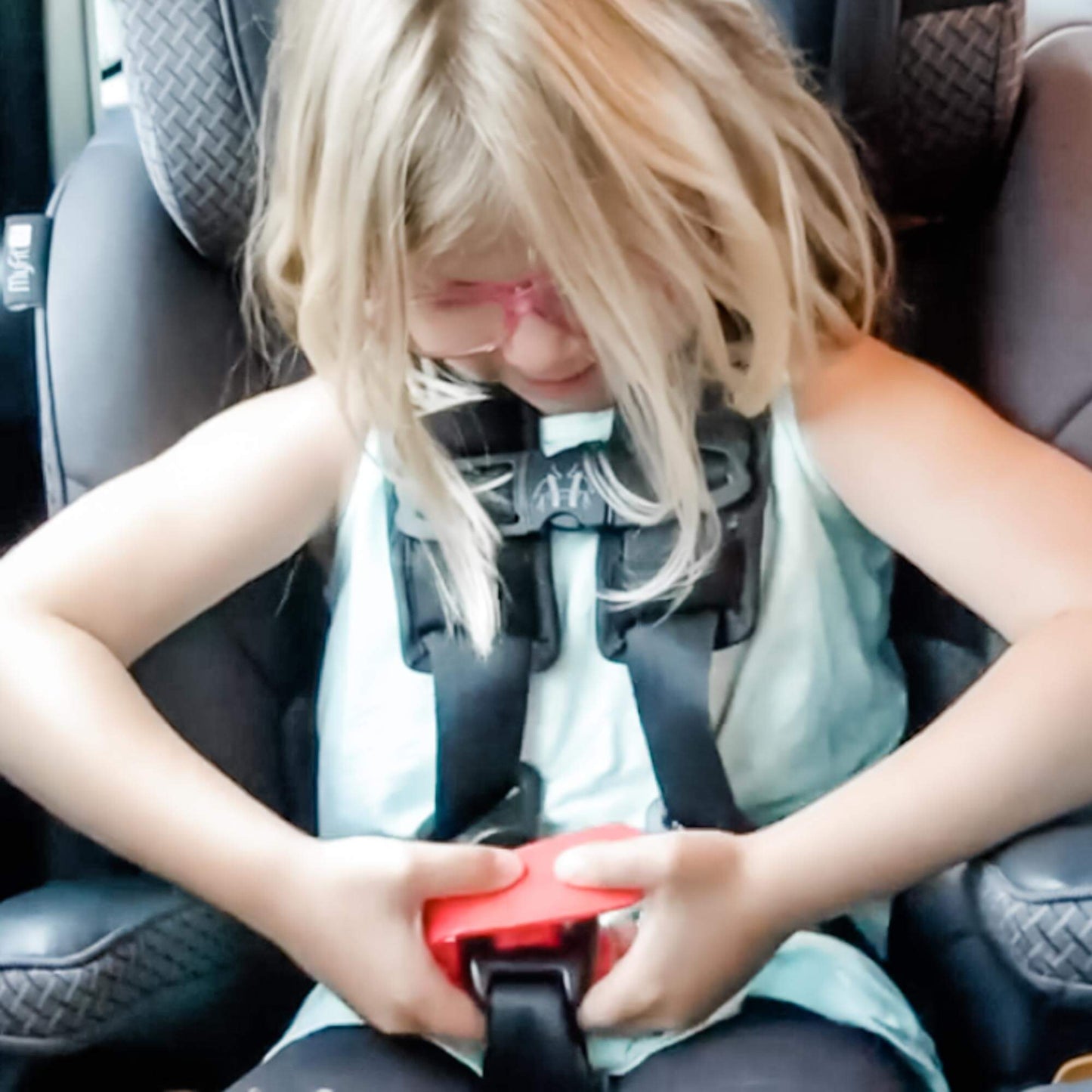 Child in a car seat being buckled up with a red strap.