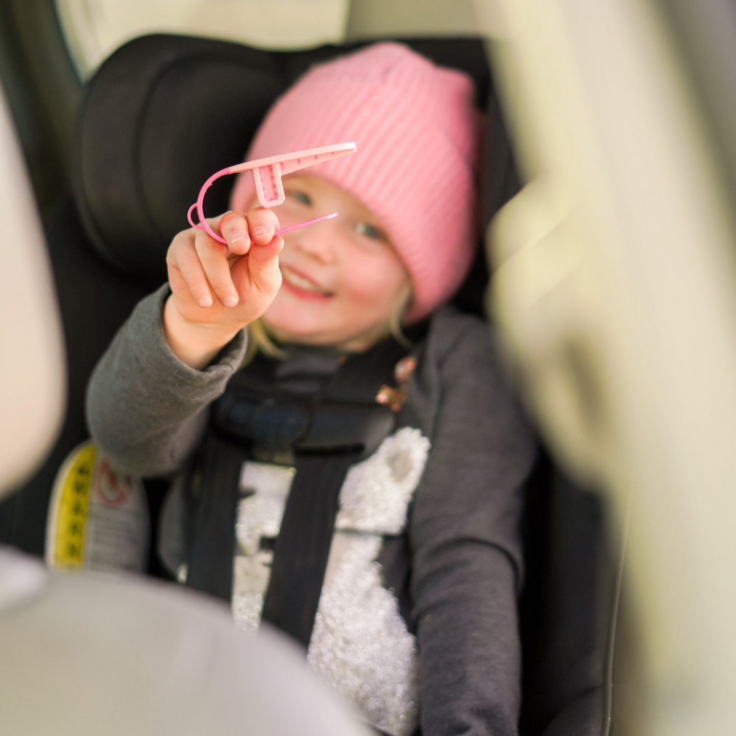 Child in a car seat holding a pink object, wearing a pink hat.