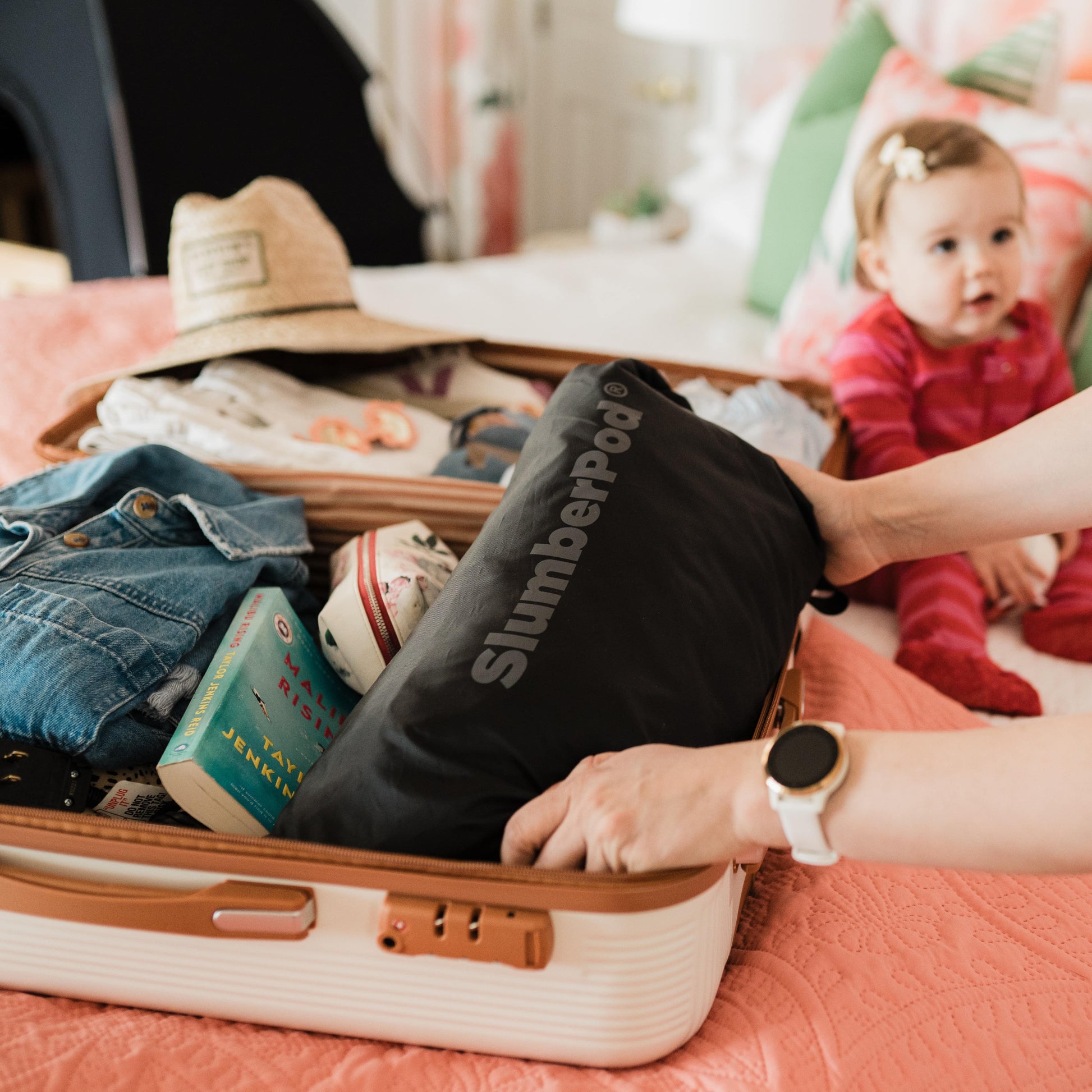 A close-up of a person's hands packing a black SlumberPod carrying bag into an open hardshell suitcase. The suitcase is partially filled with travel essentials like denim clothing and a book. In the background, a young toddler in pink striped pajamas sits on a bed, illustrating the product's convenience for family travel.