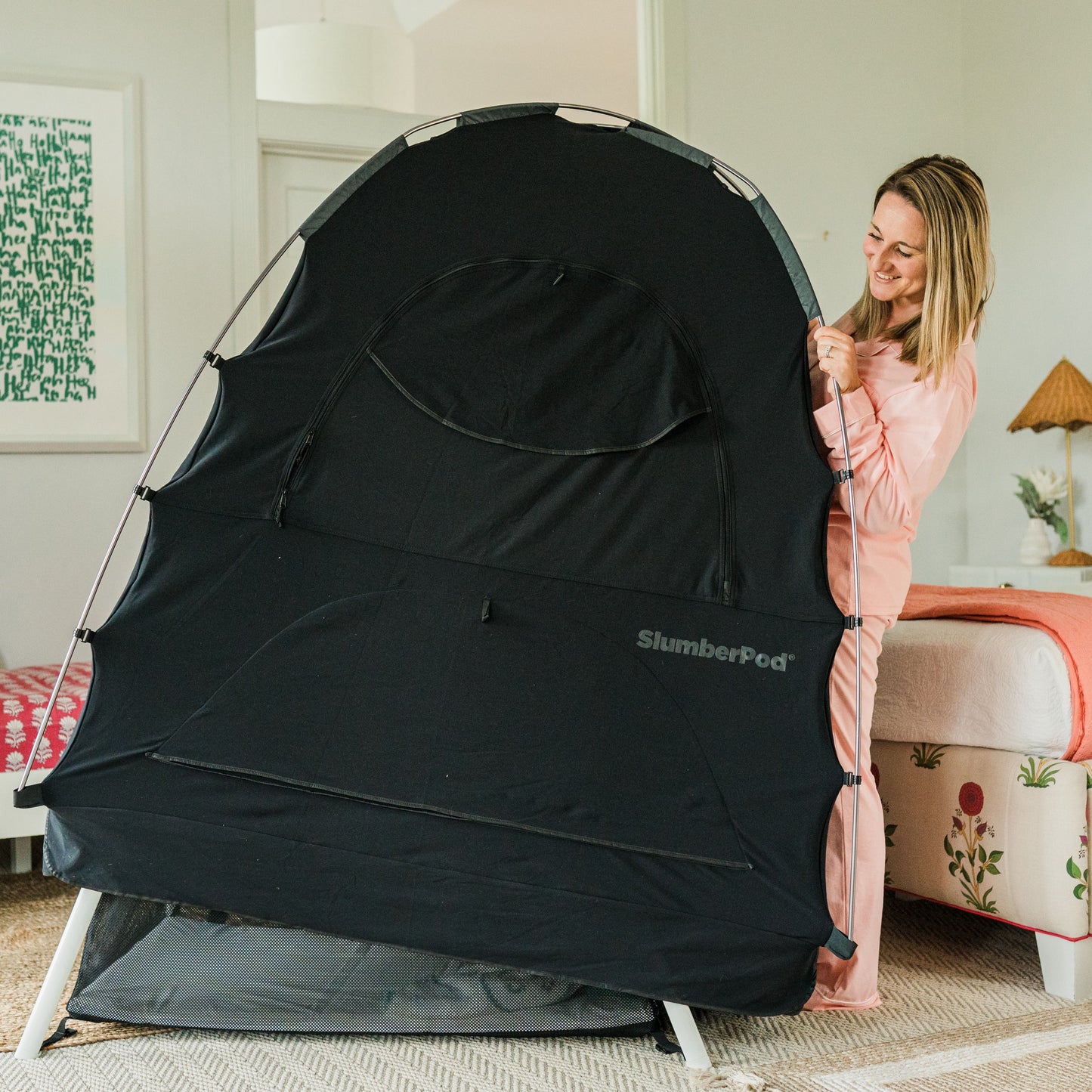 A woman in pink pajamas stands in a bright bedroom, smiling as she fits a black SlumberPod privacy tent over a white-framed travel playard. The tent is nearly fully positioned, showing its large arched shape and 'SlumberPod' branding. The room is decorated with floral-patterned furniture and a framed piece of green text art on the wall.