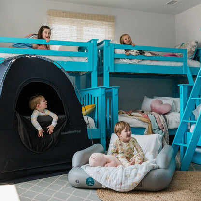 Four happy children playing in a brightly lit room with blue bunk beds. A toddler peeks out from the mesh window of a black SlumberPod privacy tent, while another young child sits comfortably on a gray inflatable travel cot in the foreground. Two older children look down smiling from the top of the blue bunk beds, showcasing a complete travel sleep setup for a large family.