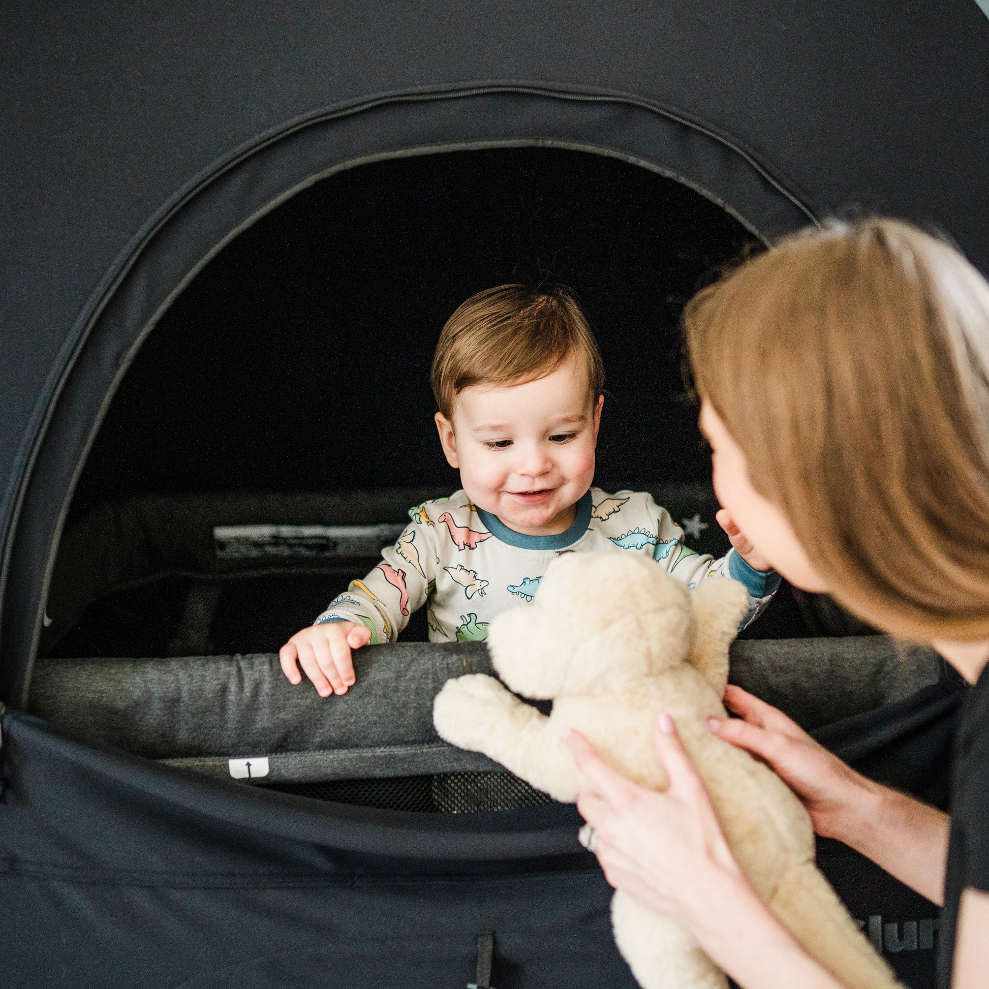 A close-up of a smiling toddler in dinosaur-patterned pajamas standing inside a black SlumberPod privacy tent. A woman in the foreground is shown from the side, handing a stuffed toy dog to the child through the tent's large zippered opening. The interior of the tent is dark, illustrating its blackout capabilities for sleep.