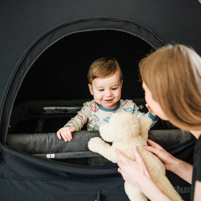 A close-up of a smiling toddler in dinosaur-patterned pajamas standing inside a black SlumberPod privacy tent. A woman in the foreground is shown from the side, handing a stuffed toy dog to the child through the tent's large zippered opening. The interior of the tent is dark, illustrating its blackout capabilities for sleep.