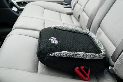 A black and grey BubbleBum inflatable travel booster seat sits ready on the light-colored leather back seat of a car. A red seatbelt positioning clip is visible at the bottom corner.