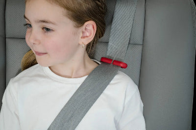 A close-up shot of a young girl with braided hair sitting in a car seat, looking off to the side. She is wearing a grey three-point seatbelt that is properly positioned across her shoulder. A small, bright red belt positioning clip is attached to the shoulder strap, ensuring the belt stays away from her neck for safety and comfort.
