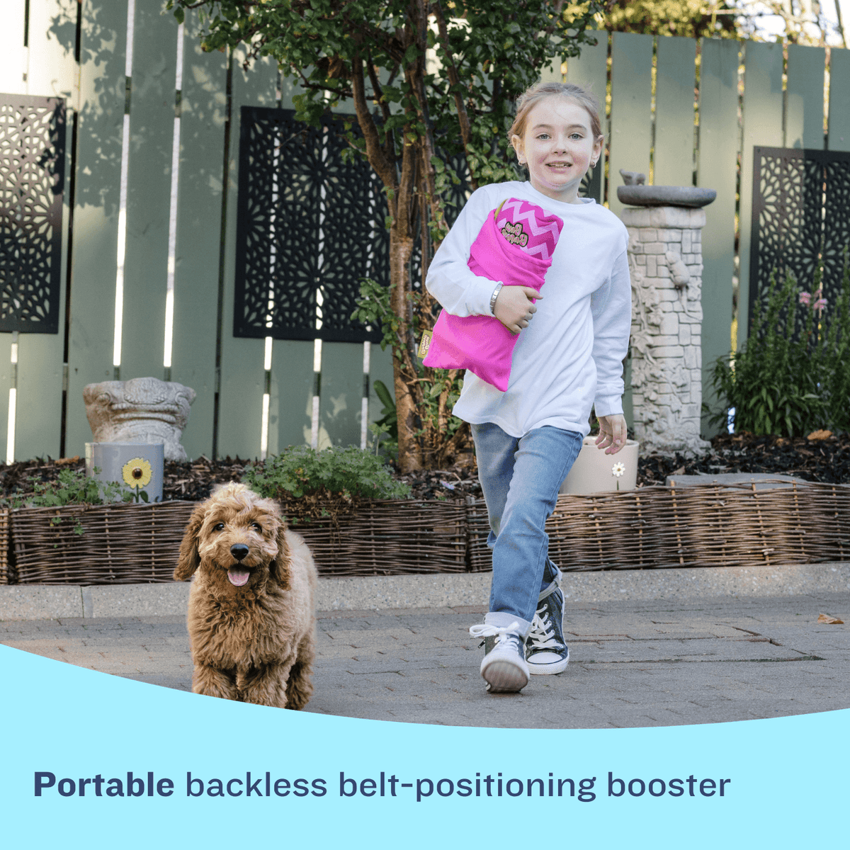 A young girl in a white shirt and jeans runs happily along a paved path with a small, fluffy brown dog. She is carrying a compact, rolled-up pink BubbleBum travel bag under her arm. A light blue graphic at the bottom contains the text: "Portable backless belt-positioning booster."
