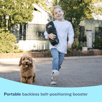 A young girl in a white shirt and jeans runs happily along a paved path with a small, fluffy brown dog. She is carrying a compact, rolled-up black BubbleBum travel bag under her arm. A light blue graphic at the bottom contains the text: "Portable backless belt-positioning booster."