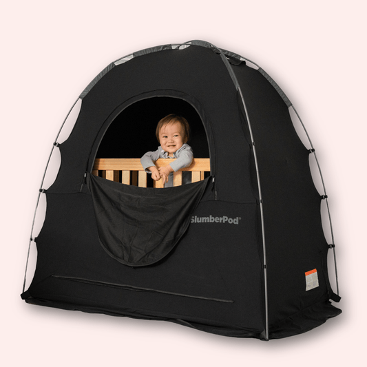 A smiling toddler sits inside a wooden mini crib that is fully enclosed by a black SlumberPod privacy tent. The toddler is looking out of the tent's large, circular ventilation window, which has its fabric flap tucked down. The tent features a sturdy arched frame and the white 'SlumberPod' logo on the side.