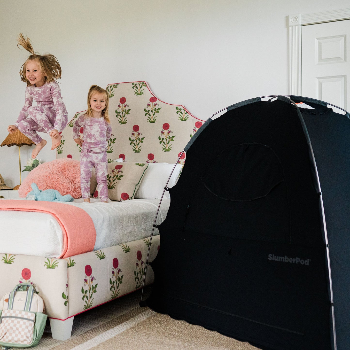 A bright bedroom scene featuring two young girls in matching purple tie-dye pajamas playing on a floral-patterned bed. One girl is jumping in the air while the other stands smiling next to a large black SlumberPod privacy tent positioned on the floor beside the bed. A small patterned backpack sits on the floor in the foreground.