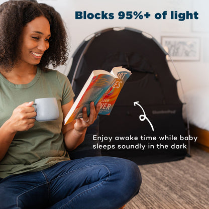 A lifestyle marketing graphic for SlumberPod featuring a woman sitting on the floor in the foreground, smiling while reading a book and holding a mug. In the background, a black SlumberPod privacy tent is set up, indicated by a white arrow. The text at the top reads 'Blocks 95%+ of light,' while text at the bottom says 'Enjoy awake time while baby sleeps soundly in the dark'.