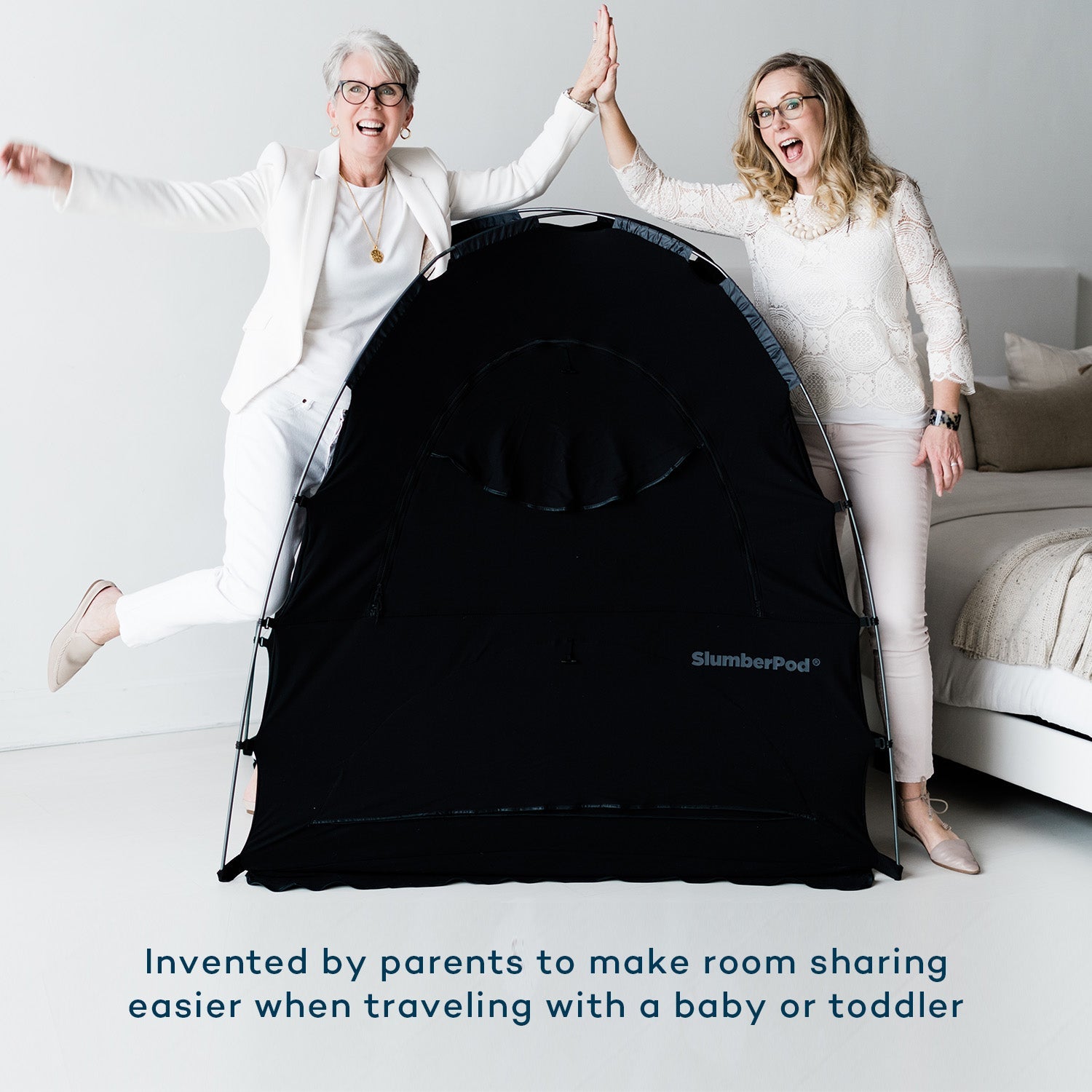 Two cheerful women, the inventors of SlumberPod, give each other a high-five while standing behind a black portable SlumberPod sleep tent. They are in a bright, modern room next to a bed with white linens. Text at the bottom of the image reads: 'Invented by parents to make room sharing easier when traveling with a baby or toddler'.