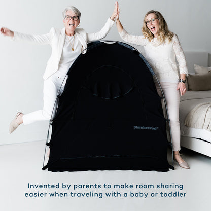 Two cheerful women, the inventors of SlumberPod, give each other a high-five while standing behind a black portable SlumberPod sleep tent. They are in a bright, modern room next to a bed with white linens. Text at the bottom of the image reads: 'Invented by parents to make room sharing easier when traveling with a baby or toddler'.