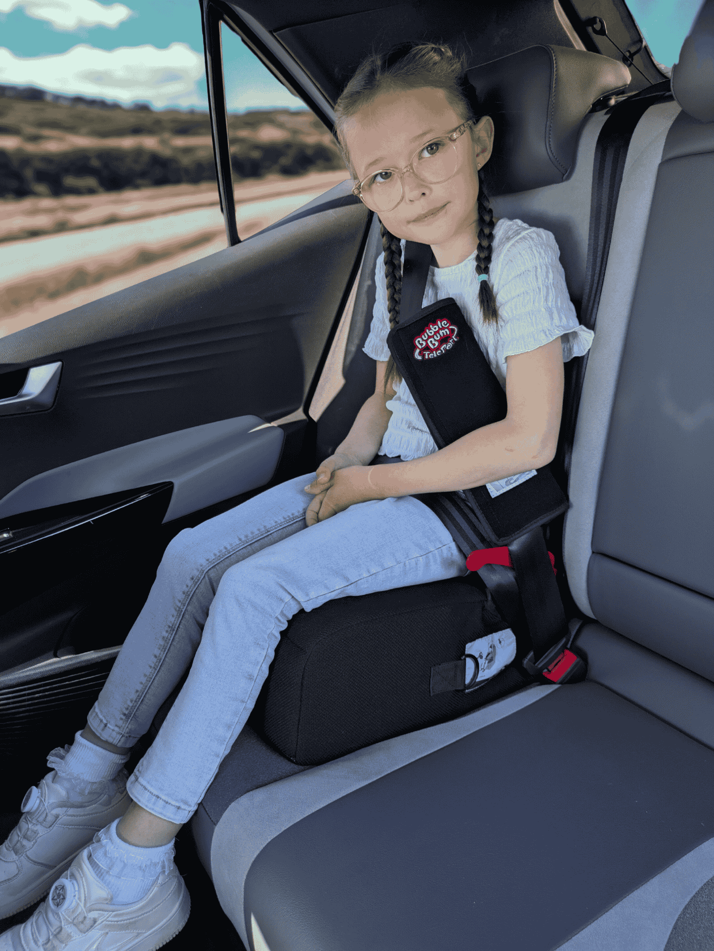 A young girl with glasses and braided hair sits safely in the back of a car using a black BubbleBum TelePort booster seat. She is wearing a white t-shirt and blue jeans. The booster seat elevates her so the seat belt fits properly across her shoulder and lap, and a black padded seat belt cover with the red BubbleBum TelePort logo is visible on the shoulder strap.