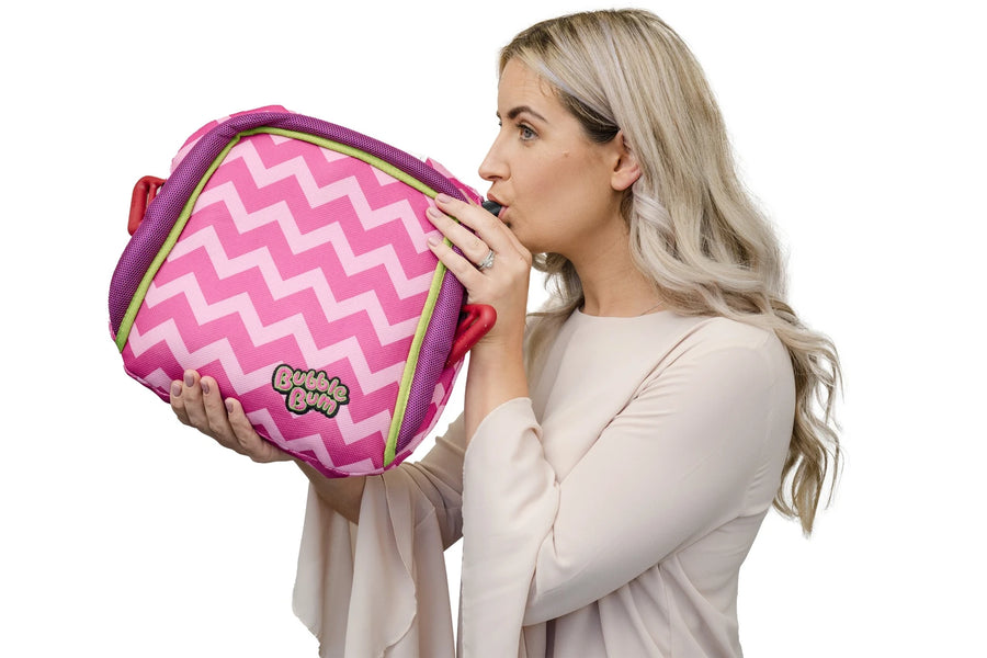 A woman with blonde hair against a white background, demonstrating how to inflate a pink and white chevron-patterned BubbleBum booster seat. She is holding the seat with both hands and blowing into the inflation valve on the side of the seat.