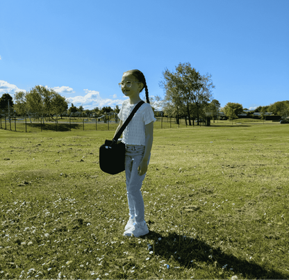 A young girl with glasses and braided hair standing in a large, sunny green field under a clear blue sky. She is wearing a white shirt and light-blue jeans, carrying a black BubbleBum booster seat in its compact shoulder-strap carrying bag. The background features a distant playground and trees.