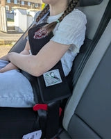 A close-up of a child sitting in a car seat wearing a black padded seatbelt adjuster. The adjuster features a red logo at the top and an instructional diagram at the bottom, helping to position the shoulder strap away from the neck.